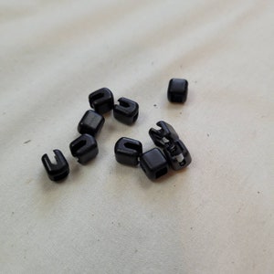 Puede incluir: Un grupo de diez cierres de cord&oacute;n de pl&aacute;stico negro, tambi&eacute;n conocidos como topes de palanca, est&aacute;n esparcidos sobre una superficie de tela blanca.