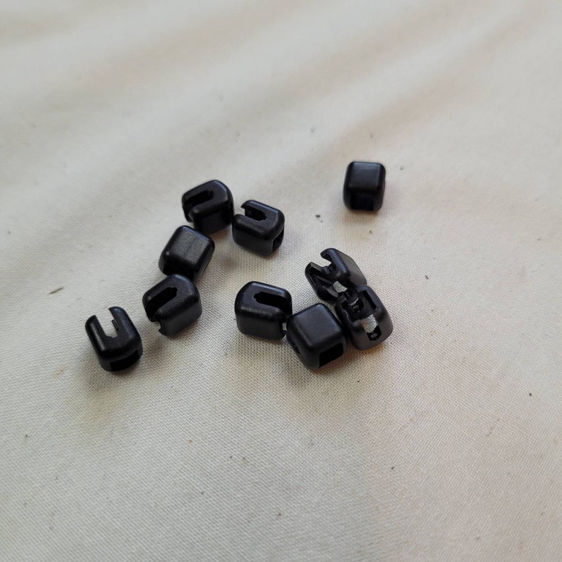 Puede incluir: Un grupo de diez cierres de cord&oacute;n de pl&aacute;stico negro, tambi&eacute;n conocidos como topes de palanca, est&aacute;n esparcidos sobre una superficie de tela blanca.