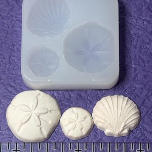 May include: A clear silicone mold with three seashell designs and two finished seashell castings. The mold features a scallop shell and two sand dollars. The finished castings are off-white and sit on a purple surface next to a ruler.