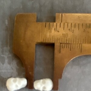 May include: Two white ceramic skull-shaped beads measured with a metal caliper. The caliper is open to approximately 1.14 inches.