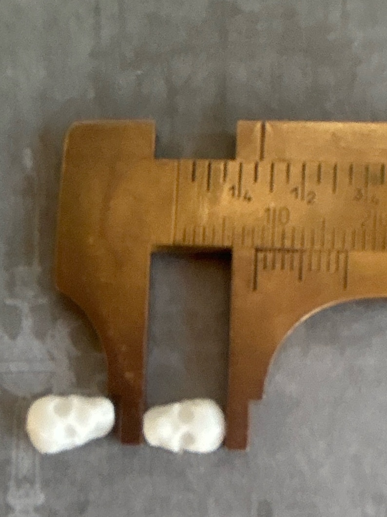 May include: Two white ceramic skull-shaped beads measured with a metal caliper. The caliper is open to approximately 1.14 inches.