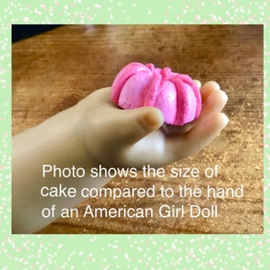 May include: A pink cake shaped like a flower, held in the hand of an American Girl doll. The text "Photo shows the size of cake compared to the hand of an American Girl Doll" is visible below the hand.