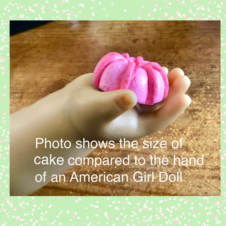 May include: A pink cake shaped like a flower, held in the hand of an American Girl doll. The text "Photo shows the size of cake compared to the hand of an American Girl Doll" is visible below the hand.
