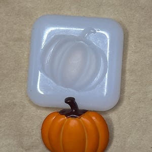 May include: An orange pumpkin figurine with a dark brown stem next to a clear, square-shaped silicone mold in the shape of a pumpkin. The pumpkin is a fall-themed decorative item.