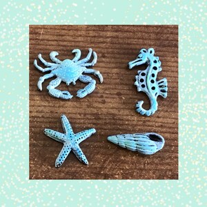 May include: Four light blue metal sea-themed charms: a crab, seahorse, starfish, and seashell. Each charm has a distressed finish, with black accents. The charms are arranged on a brown wooden surface.