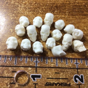 May include: A collection of small, white skull-shaped objects scattered on a brown surface. A clear ruler with black markings and the word "FISKARS" is in the foreground, along with a ring. The skulls appear to be made of a solid material.