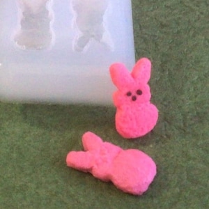 May include: Two pink bunny-shaped candies, one with a slightly different shape than the other, are shown on a green surface. The candies are made in a silicone mold, which is also visible in the image.