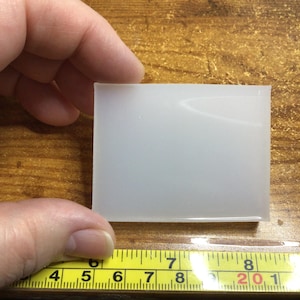 May include: A translucent, rectangular object is held against a wooden surface. The object appears to be approximately 3 inches wide. A yellow measuring tape with black markings is visible in the foreground, indicating the object's dimensions.