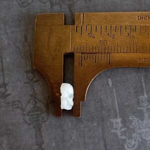 May include: A white ceramic skull charm measured with a brass ruler. The skull is approximately 1/2 inch in size.