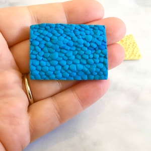 May include: A close-up of a blue polymer clay sheet with a textured pattern of scales. The sheet is being held in a hand.