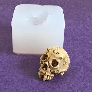 May include: A small, detailed skull figurine in a light beige color sits next to a white, rectangular mold. The skull has visible eye sockets, a nose cavity, and teeth. The mold is likely for creating similar skull-shaped items.