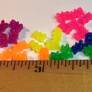 May include: A collection of colorful gummy bear candies in various colors, including pink, purple, orange, yellow, green, and blue. A wooden ruler with inch markings is in the foreground, providing scale.
