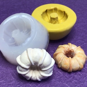 May include: A collection of resin molds and finished castings. The molds are a clear, translucent silicone and bright yellow. The castings are white and tan, shaped like a flower or a decorative ring. The background is a purple fabric.