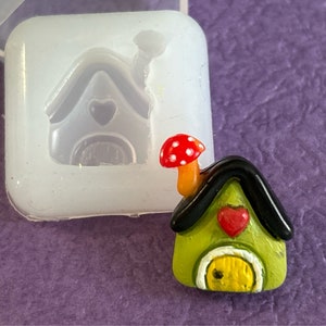 May include: A small green house with a black roof and a red heart on the front. The house has a yellow door and a red and white mushroom on top. The house is made of polymer clay and is about 1 inch tall.