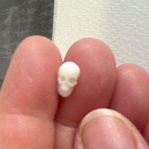 May include: A small white skull charm, held in a hand. The skull has a detailed design with eye sockets and a nose.