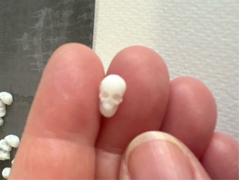 May include: A small white skull charm, held in a hand. The skull has a detailed design with eye sockets and a nose.