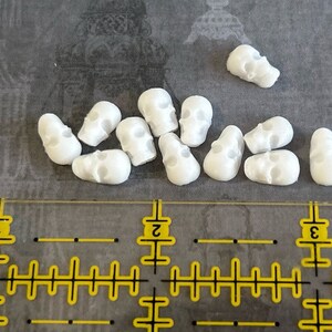 May include: A collection of 12 small white skull beads. The beads are made of a smooth, white material and have a realistic skull design.
