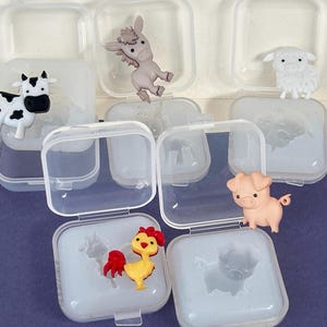 May include: A collection of small, plastic farm animal figurines, including a cow, donkey, sheep, pig, rooster, and pig, each displayed in a clear, square container with a hinged lid. The figurines are brightly colored and detailed.