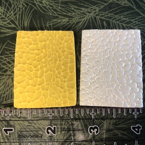 May include: Two rectangular, textured objects. One is yellow, the other is white. Both have a pattern of small, irregular shapes. A ruler with inch markings is at the bottom, with the objects resting on a green patterned surface.