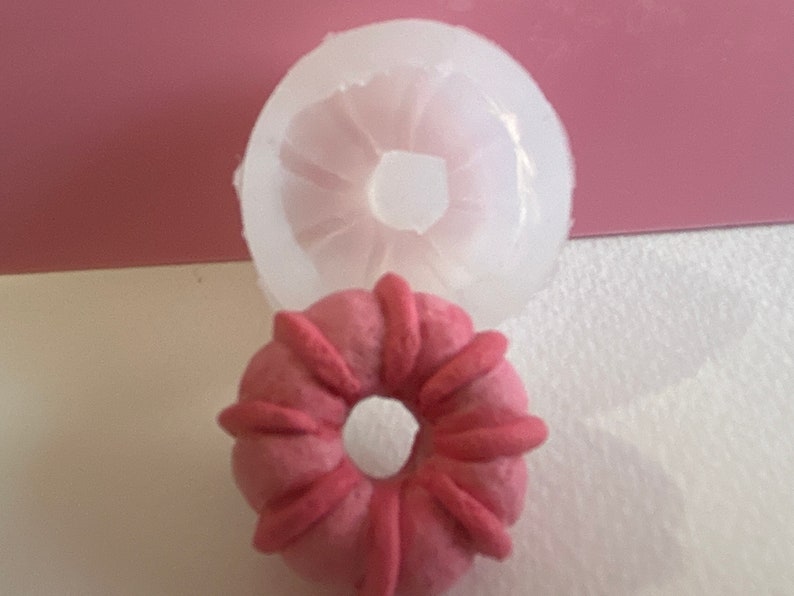 May include: A pink silicone mould for making donut-shaped pastries. The mould is shown with a pink donut-shaped pastry inside.