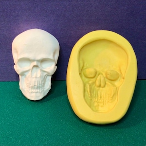 May include: A white skull figurine next to a yellow skull-shaped mold. The skull has detailed facial features, including eye sockets, a nose cavity, and teeth. The mold is a negative impression of the skull.