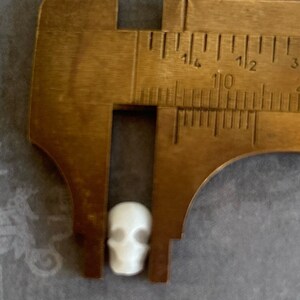 May include: A white ceramic skull bead measures approximately 1 cm in diameter. The bead is being measured with a metal caliper.