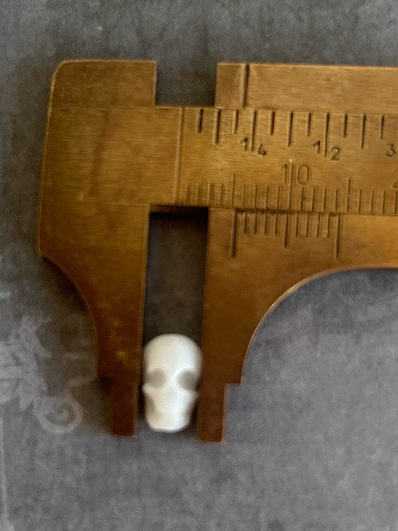 May include: A white ceramic skull bead measures approximately 1 cm in diameter. The bead is being measured with a metal caliper.