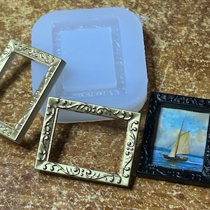 May include: A collection of miniature frames and a silicone mold. Two gold-colored, ornate frames sit beside a black frame with a small painting of a sailboat. A clear, square silicone mold with a frame design is also present.