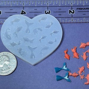 May include: A heart-shaped translucent silicone mold featuring cavities for fish and tail shapes, positioned next to a ruler showing approximately 3.5 inches in width and a US quarter coin, with several small orange fish and blue tail charms.