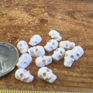 May include: A collection of 12 small white ceramic skull charms, scattered on a brown wooden surface. The skulls are approximately the size of a US quarter.