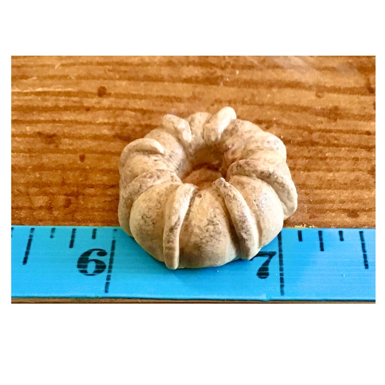 May include: A small, light brown, ring-shaped cake with a textured surface. The cake is approximately 2.5 cm in diameter and is resting on a blue measuring tape.