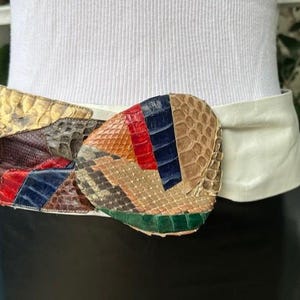 May include: A wide, cream-coloured leather belt with a large, oval buckle. The buckle is made of various patches of snakeskin in shades of gold, brown, red, blue, and green. The belt is worn around the waist.