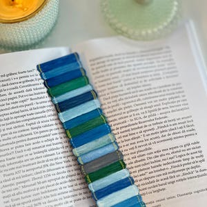 Könnte beinhalten: Handgefertigter Lesezeichen mit farbenfrohem Design. Das Lesezeichen hat ein Muster aus blauen, grünen und grauen Fäden, ideal zum Markieren von Seiten in einem Buch. Das Lesezeichen liegt auf einem offenen Buch.