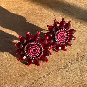 Puede incluir: Un par de pendientes de cuentas rojas con un centro de tela burdeos. Los pendientes tienen forma de flor con un diseño en espiral en el centro.