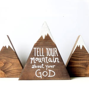 Könnte beinhalten: Drei dreieckige Holzberge mit weiß bemalten Schneegipfeln. Der mittlere Berg trägt die Worte "TELL YOUR MOUNTAIN ABOUT YOUR GOD" in weißer Farbe.