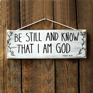 Może przedstawiać: Biały drewniany znak z przetartym wykończeniem, wiszący na białym sznurku. Znak ma czarny tekst, który brzmi "Be still and know that I am God" z odniesieniem do Pisma Świętego "Psalm 46:10" poniżej. Znak jest ozdobiony zielonymi, liściastymi winoroślami po obu stronach.