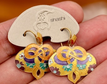 Può includere: Orecchini dorati con un design smaltato colorato. Gli orecchini presentano un motivo floreale e astratto con accenti viola, verdi e blu. Gli orecchini sono su una carta beige con il marchio "shashi".