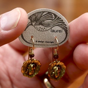 Pode incluir: Brincos de ouro com um design floral e pequenas contas marrons penduradas na parte inferior. Os brincos estão em um cartão com a palavra "laurel" e o texto "design copyright".