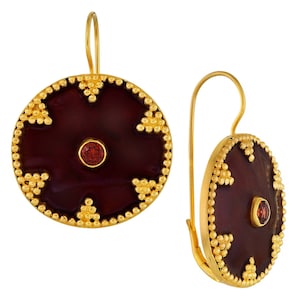 May include: A pair of gold earrings with a round, maroon-colored stone set in the center of each earring. The stone is surrounded by a gold design of small circles and triangles. The earrings have a hook closure.