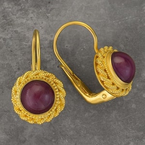 Peut inclure: Une paire de boucles d'oreilles en or avec une pierre précieuse en cabochon rubis sertie dans un design de fil torsadé. Les boucles d'oreilles ont une fermeture à levier.