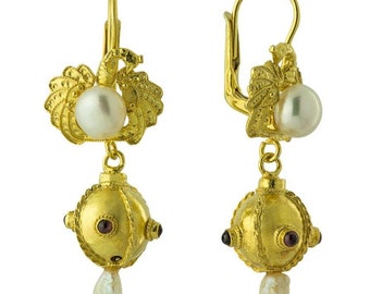 Puede incluir: Pendientes de oro con una perla y una pequeña bola adornada con piedras preciosas rojas. Los pendientes tienen un diseño delicado e intrincado.