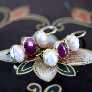 May include: A pair of gold earrings featuring a white pearl and a purple gemstone. The earrings are drop earrings with a hook fastening.