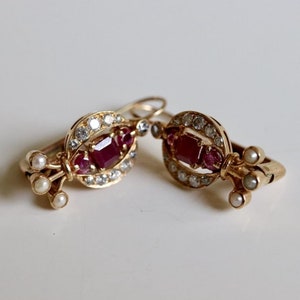 May include: A pair of gold earrings with ruby and diamond accents. The earrings feature a cluster of pearls on the bottom.