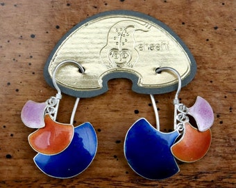 May include: A pair of colorful dangle earrings with a silver hook. Each earring features three enamel shapes in blue, orange, and pink. The earrings hang from a gold-colored tag with a monkey design and the word "sheski".