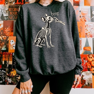May include: A dark charcoal grey crewneck sweatshirt featuring a graphic print of a dog skeleton being touched on the nose by a skeletal finger. The word boop is printed in a white script font above the dog's head, centred on the chest.