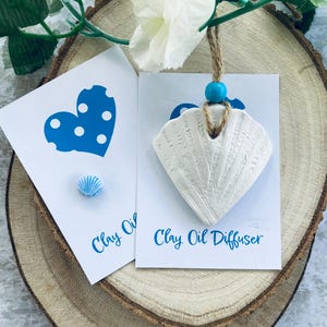 May include: A white clay shell-shaped oil diffuser with a turquoise bead and jute twine hanger, displayed on a white card with the text Clay Oil Diffuser. A smaller blue and white shell-shaped charm is on a separate card with a blue heart graphic.