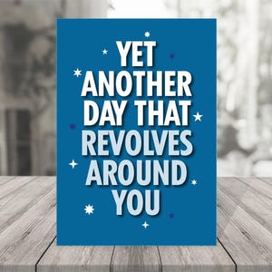 May include: A blue greeting card with white text that reads "YET ANOTHER DAY THAT REVOLVES AROUND YOU." The card is decorated with small white stars and blue dots. The card is on a wooden surface.