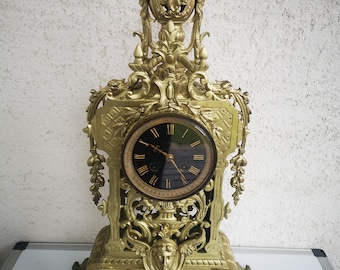 May include: Ornate gold-colored mantel clock with a black clock face and Roman numerals. The clock features intricate decorative details, including floral and face motifs, and a decorative top. The clock is set on a rectangular base.