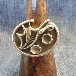 May include: A silver ring with an oval face featuring an abstract design. The ring's face has a central, stylized element with curved lines and two small, flower-like details. The ring is displayed on a wooden ring holder against a denim background.