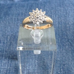 Könnte beinhalten: Ein goldener Ring mit einem großen, runden, klaren Diamanten-Cluster in der Mitte. Der Ring befindet sich auf einem transparenten Präsentationsständer.